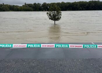 Zaplavené nábrežie rieky s policajnou páskou „Vstup zakázaný“, pričom voda vystúpila až k stromom.