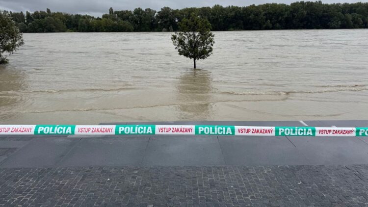 Zaplavené nábrežie rieky s policajnou páskou „Vstup zakázaný“, pričom voda vystúpila až k stromom.