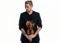 David Garrett drží husle v rukách, oblečený v čiernom saku, s výrazom sústredenia, na svetlom pozadí.