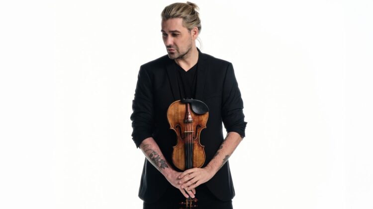 David Garrett drží husle v rukách, oblečený v čiernom saku, s výrazom sústredenia, na svetlom pozadí.