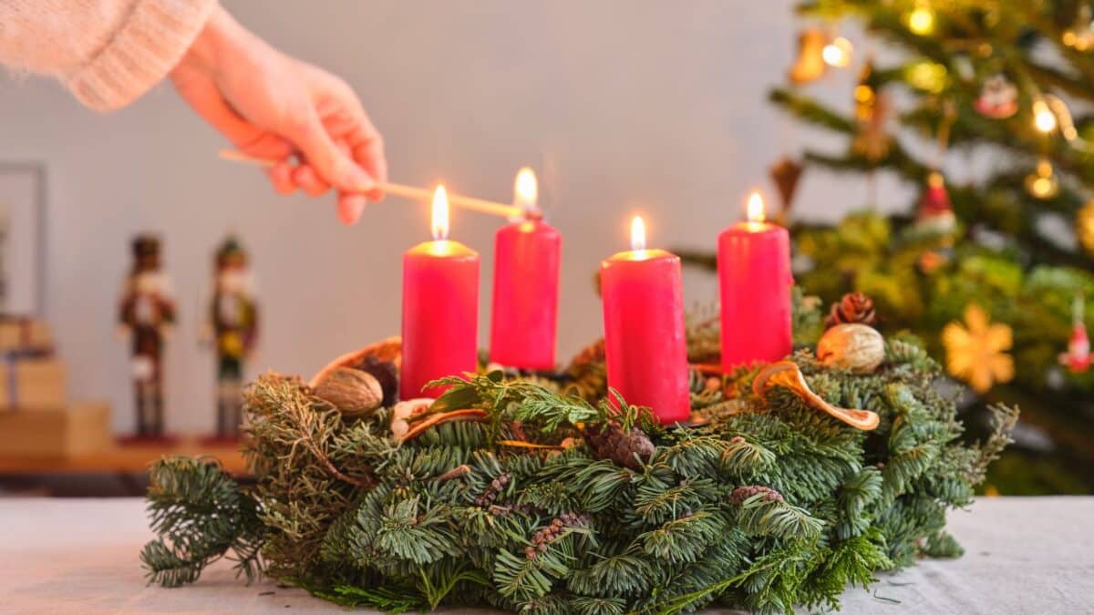 Adventný veniec s červenými sviečkami, z ktorých jedna sa zapaľuje zápalkou. V pozadí je vianočný stromček a drevené dekorácie.