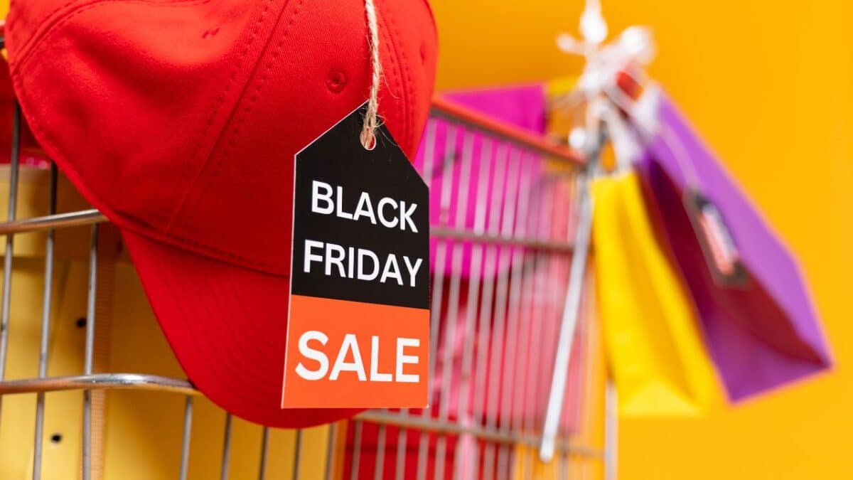 Červená šiltovka s visačkou "Black Friday Sale" na nákupnom košíku, obklopenom farebnými taškami na žltom pozadí