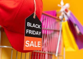 Červená šiltovka s visačkou "Black Friday Sale" na nákupnom košíku, obklopenom farebnými taškami na žltom pozadí