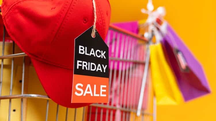 Červená šiltovka s visačkou "Black Friday Sale" na nákupnom košíku, obklopenom farebnými taškami na žltom pozadí