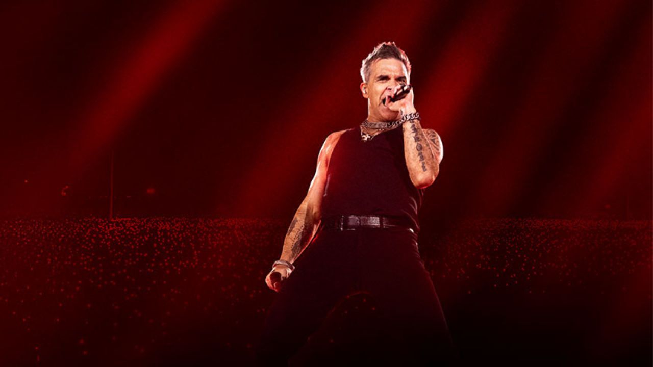 Robbie Williams spieva na pódiu počas koncertu, v čiernom oblečení a s mikrofónom v ruke, osvetlený červenými svetlami.