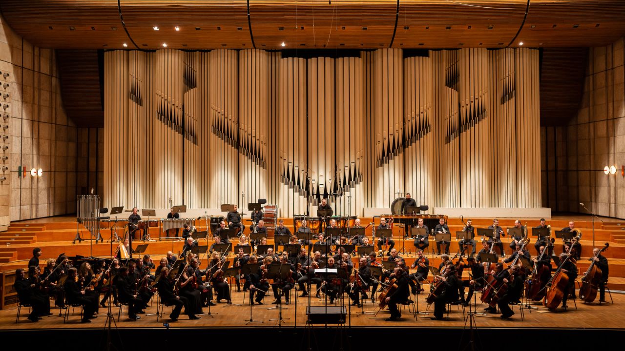 Symfonický orchester vystupuje na javisku pred veľkými organovými píšťalami v koncertnej sále.