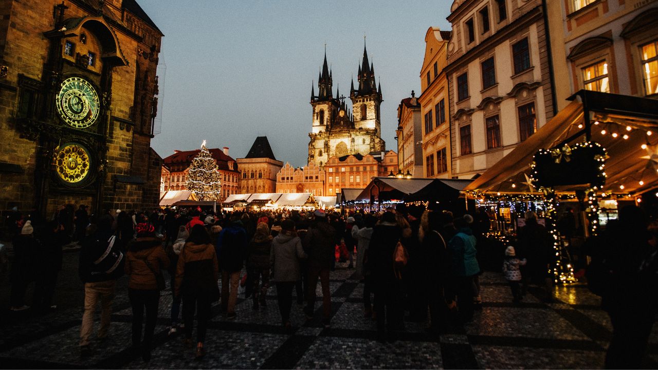 Námestie s historickými budovami, vianočnými trhmi a osvetleným vianočným stromčekom počas zimného večera. Dav ľudí si užíva sviatočnú atmosféru a stánky ozdobené svetielkami.