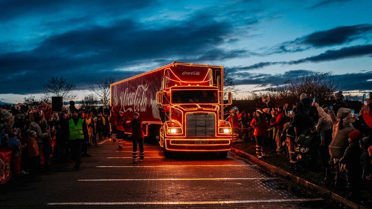 Vianočný Coca Cola kamión ozdobený vianočnými svetlami, obklopený davom ľudí v podvečernom svetle, prichádza na miesto podujatia.