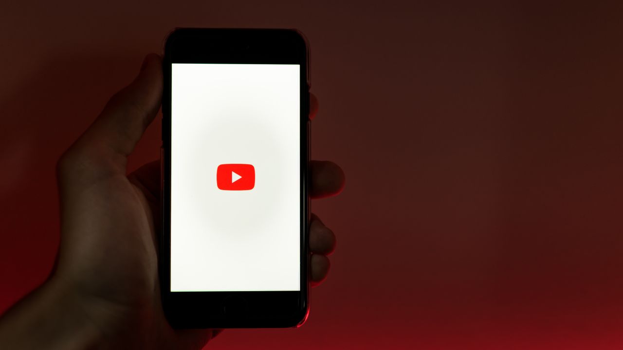 Ruka držiaca mobilný telefón s logom YouTube na obrazovke, s tmavým pozadím.