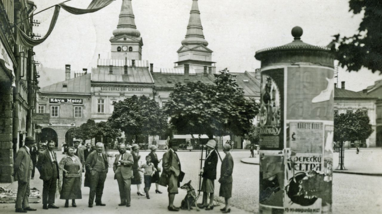 Historická fotografia Žiliny z medzivojnového obdobia, zachytávajúca skupinku ľudí na námestí s dvojicou veží v pozadí.