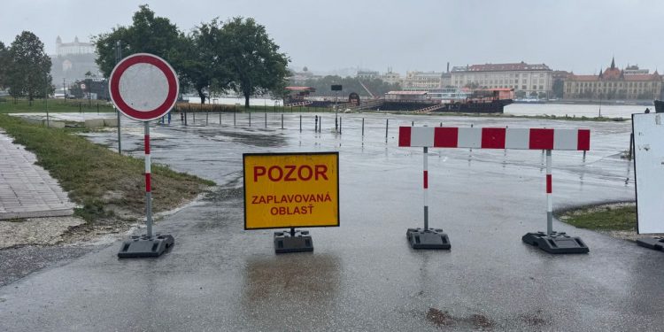 Záplavová oblasť označená dopravnou značkou a varovným žltým nápisom 'Pozor: zaplavovaná oblasť,' s pohľadom na zaplavenú cestu a Bratislavský hrad v pozadí.