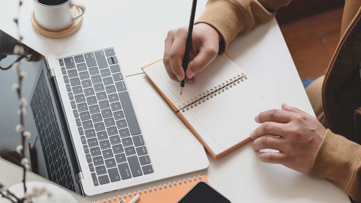 Osoba si robí poznámky do zápisníka pri notebooku a šálke kávy.