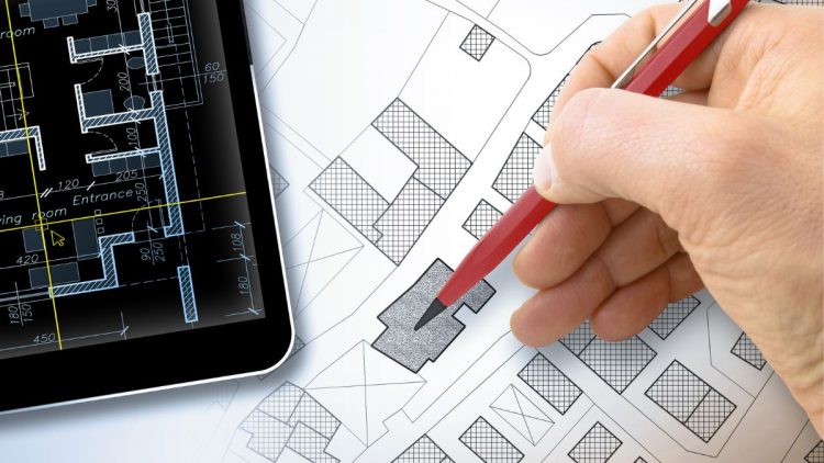 Ruka drží červenú ceruzku nad katastrálnou mapou, vedľa tabletu zobrazujúceho stavebné plány.