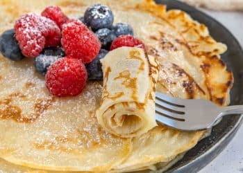 Detailný pohľad na zlatisté palacinky s čerstvými malinami a čučoriedkami, posypané práškovým cukrom, podávané na čiernom tanieri.