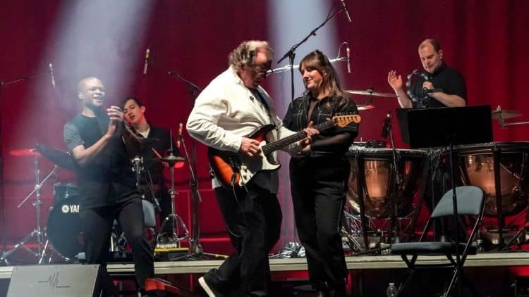 Hudobníci na pódiu počas koncertu Mike Oldfield’s Tubular Bells in Concert. Dirigent Robin Smith hrá na elektrickej gitare obklopený ďalšími členmi kapely v atmosférickom osvetlení.