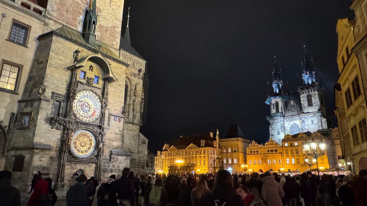 Nočná atmosféra Staromestského námestia v Prahe s osvetleným Pražským orlojom a Kostolom Matky Božej pred Týnom, pred ktorými sa nachádza dav turistov.